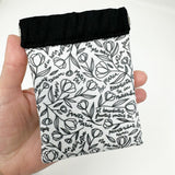 Squeeze pouch measuring approximately 5" tall x 4" wide. Outer fabric is a black and white floral print with Latin text of the Hail Mary dispersed throughout. Inside accent is a black fabric. Snap frame for easy opening and closing.