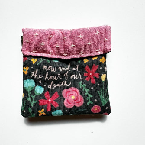 Clasp pouch measuring approximately 3.75” wide x 3.5” wall x 1” deep. Outer fabric is a bright and cheery black floral background with the words of the Hail Mary interspersed throughout. Inside lining is a pink fabric with cross stitch embroidery design. Clasp frame for easy opening and closing.