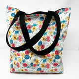 Lined tote bag measuring approximately 12" wide x 14" tall x 2.5" deep. Exterior fabric features white background with bright watercolor flowers and a hidden rosary design. Black lining. Black cotton webbing straps.