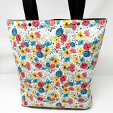 Lined tote bag measuring approximately 12" wide x 14" tall x 2.5" deep. Exterior fabric features white background with bright watercolor flowers and a hidden rosary design. Black lining. Black cotton webbing straps.
