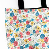 Lined tote bag measuring approximately 12" wide x 14" tall x 2.5" deep. Exterior fabric features white background with bright watercolor flowers and a hidden rosary design. Black lining. Black cotton webbing straps.