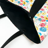 Lined tote bag measuring approximately 12" wide x 14" tall x 2.5" deep. Exterior fabric features white background with bright watercolor flowers and a hidden rosary design. Black lining. Black cotton webbing straps.