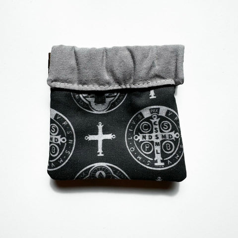 Clasp pouch measuring approximately 3.75” wide x 3.5” wall x 1” deep. Outer fabric is a black background with images of a cross and both sides of the St. Benedict medal interspersed throughout. Inside lining is a light gray fabric. Clasp frame for easy opening and closing.