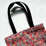 Unlined tote bag measuring approximately 13” wide x 15” tall. Exterior fabric features bright and cheery pink floral background with the words of the Hail Mary interspersed throughout. Black cotton webbing straps.
