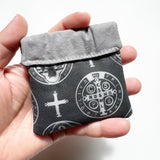 Clasp pouch measuring approximately 3.75” wide x 3.5” wall x 1” deep. Outer fabric is a black background with images of a cross and both sides of the St. Benedict medal interspersed throughout. Inside lining is a light gray fabric. Clasp frame for easy opening and closing.