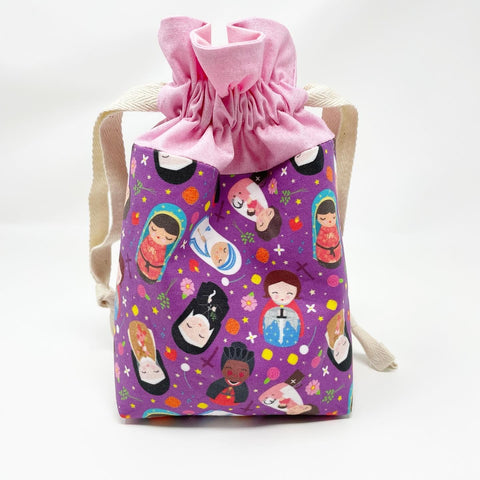 Female saints drawstring pouch; exterior main fabric has various female saints pictured; pink exterior accent piece; pink lining.